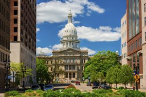 Lansing, United States - May 24, 2014 - The Michigan State Capitol as viewed from within Downtown Lansing, with trees, plants, office buildings, and pedestrians and cars and a driver in the foreground, and a blue sky with clouds in the background.